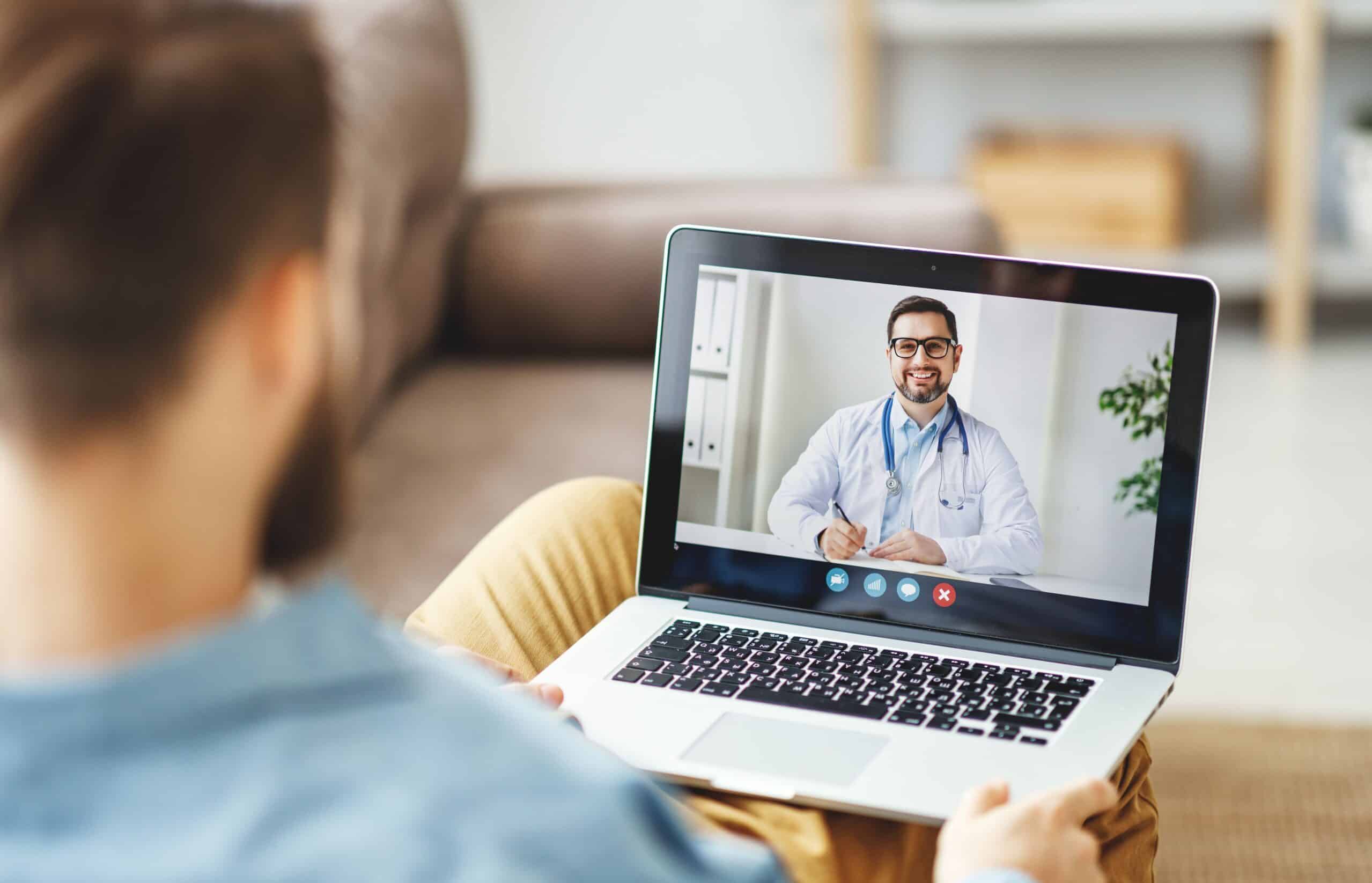 Person seated with a laptop, on a video call with a doctor wearing a stethoscope in a bright living room