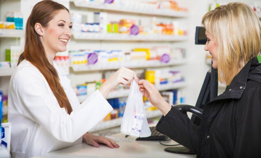 Pharmacist hands a small bag to a customer at a pharmacy counter, smiling in a well-lit aisle of products behind them.